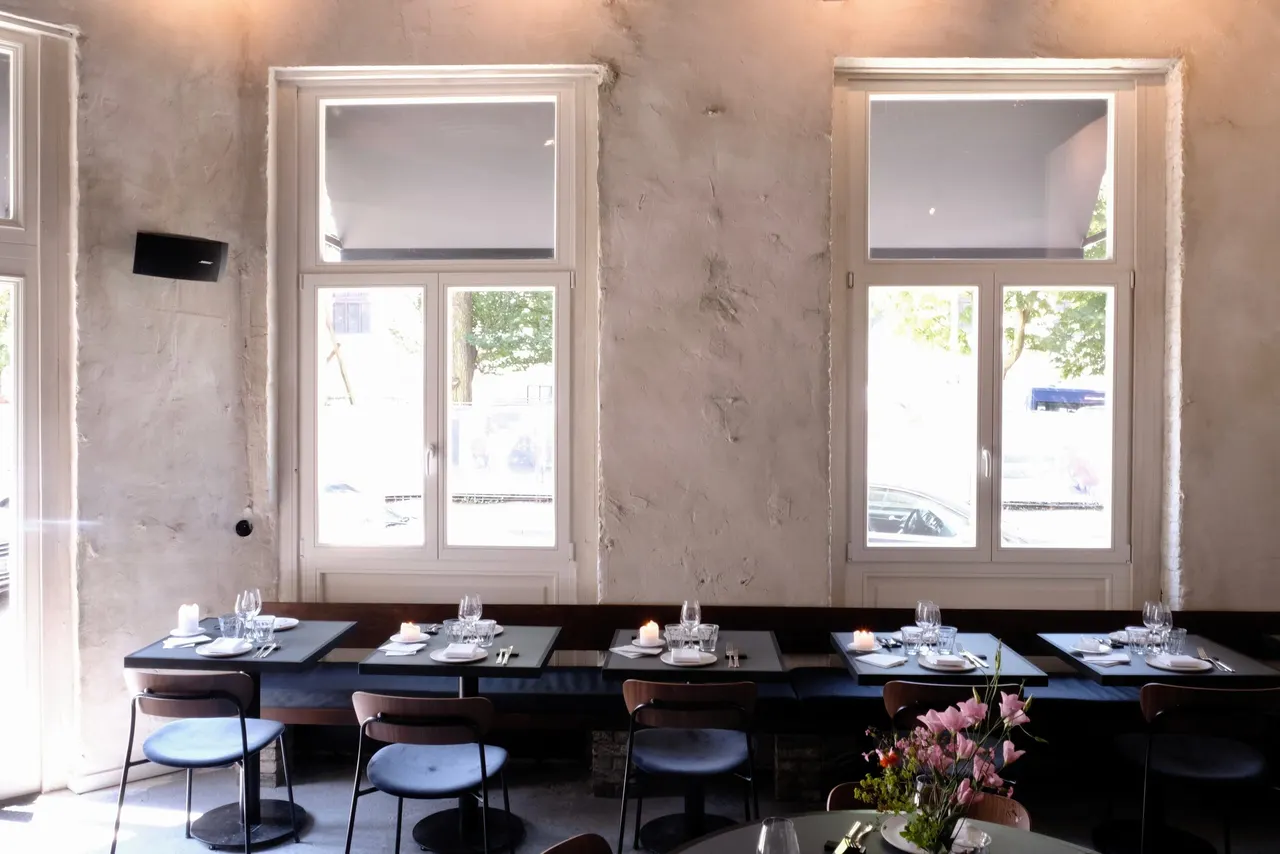 Twee hoge ramen boven een rij tafels met zwarte stoelen